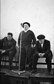 Photographie d’un homme portant un béret basque, debout face à un micro, entouré de deux hommes assis.