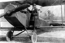 Photographie d'une femme en tenue d'aviateur debout sur une roue d'un biplan à l'arrêt.