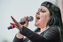 Photographie couleur d'une femme qui chante dans un micro et ferme les yeux.