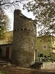 Photo prise en sous-bois. Le pigeonnier est une tour construite en moellons apparents et à toit incliné, avec un rang de carreaux émaillés de couleurs qui en fait le tour à 4 m de haut.