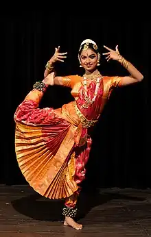 Sur fond noir, une jeune femme, portant une robe rouge et orange et de nombreux bijoux, écarte ses deux bras et lève son pied droit jusqu'à son bras droit.