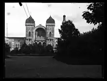 Vue en noir et blanc d'une arche encadrée de deux tours, en arrière-plan d'une haie touffue.