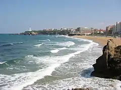 Vue d'une plage sur laquelle les vagues se brisent ; phare et bâtiments en arrière-plan.