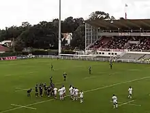 Deux équipes de rugby se font face-à-face avant de former une mêlée. En arrière-plan, la tribune d'un stade.