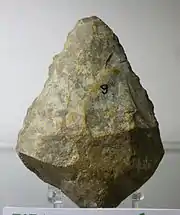 Photographie en couleurs sur fond gris d'un silex ocre, jaune et gris taillé en forme de losange irrégulier.