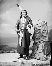 Photographie en noir et blanc d'un Amérindien en habit traditionnel coiffé d'une plume et tenant un tomahawk.