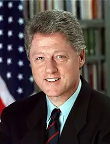 Photographie officielle du président des États-Unis, Bill Clinton. Il se tient devant le drapeau américain, porte une cravate rouge et bleue et un costume noir.