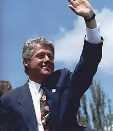 Photographie de l'ancien président américain Bill Clinton. Il porte une veste bleue, une chemise blanche et une cravate fleurie. Il fait un salut de la main gauche.