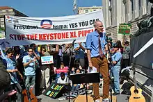 Dans une rue, un homme parle au micro sur une estrade improvisée. Derrière lui, se trouvent de nombreux manifestants, assis et debout, tenant des pancartes demandant notamment à Barack Obama de stopper la construction de l'oléoduc.