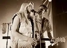 Photo d'une femme blonde qui chante à un micro avec un homme qui joue de la guitare.