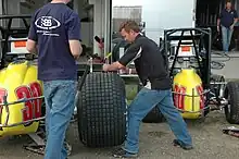 Photographie de deux hommes. Celui de gauche est de dos. Celui de droite est de profil préparant un pneu d'une voiture jaune en arrière-plan.