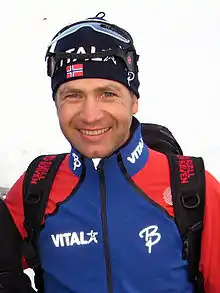 Photographie d'un homme en survêtement bleu foncé et rouge, portant un bonnet noir et des lunettes de ski relevées sur son crâne.