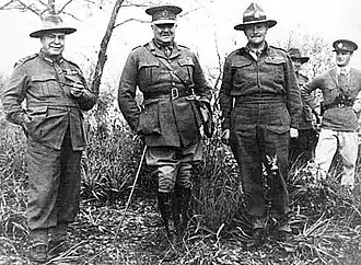 Trois officiers debout posant pour une photograhie, avec deux autres soldats au fond à droite.