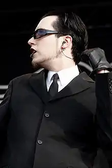 Un homme en costume noir, avec des gants noirs, du rouge à lèvres noir, des cheveux mi-longs et des lunettes noires regarde à sa droite