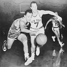Photographie en noir et blanc d'un joueur dribblant un ballon de basket-ball en courant. Il est poursuivre par un adversaire qui porte un maillot sur lequel est écrit New York.