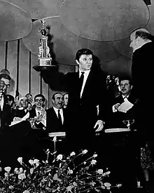 Photographie en noir et blanc d'un jeune homme debout, brandissant de la main droite un trophée : sur la droite, un homme ; au fond une foule de personnages ; devant, un grand bouquet de fleurs