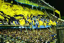 Photographie en couleurs. Dans les tribunes d'un stade, de nombreux supporters agitent des banderoles de couleur jaune et bleue.