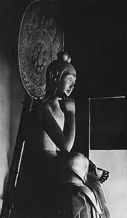 Vue de trois-quarts d'une statue assise en demi-lotus. Le pied droit repose sur la partie supérieure de la jambe gauche, le coude droit repose sur le genou droit avec la main droite près de la tête. Les cheveux de la statue sont sculptés avec deux chignons. Il y a une auréole derrière la tête de la statue.