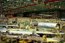 Hall d'assemblage d'avions, montrant des machines lourdes. Les ailes et les grandes sections cylindriques de l'avion sont prêtes pour l'assemblage avec d'autres composants. Au-dessus se trouvent les grues qui transportent les gros morceaux du 747.