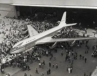 photo noir et blanc. L'avion sort d'un hangar, quelques centaines de personnes sont présentes.