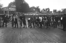 Photographie en noir et blanc d'un groupe de coureurs avant le départ, sur une piste en bois.