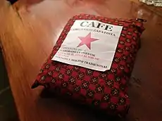 Sachet de café en tissu rouge à motif floral posé sur une table. Sur l'étiquette est inscrit en espagnol : "Café zapatiste bio. Contient 250 g. Fabriqué à Oventik, Caracol II, Altos de Chiapas. Torréfaction et broyage traditionnel." L'étiquette est décorée d'une étoile rouge au centre.