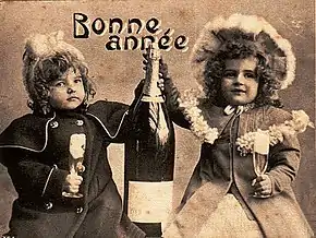 Représentation d'une carte de vœux ancienne, en noir & blanc, représentant deux enfants tenant une bouteille de champagne entre eux. Entre leurs têtes et au-dessus de la bouteille, la mention « Bonne année ».