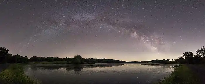 Sur un lac et dans un ciel noir, seulement éclairé par quelques ombres venues de derrière l'horizon, un arc d'étoiles se dessine au firmament.