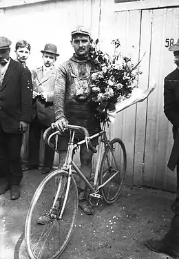 Photographie noir et blanc d'un cycliste debout sur son vélo, portant un bouquet.