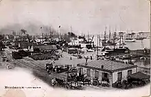 Photo sépia montrant des quais avec bâtiments bas, personnages, carrioles, tonneaux et, au fond, de grands voiliers sur l'eau