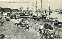 Ancienne photographie des quais de Bordeaux. Plusieurs véhicules sont tractés par des chevaux. De nombreux bateaux sont amarés au bord de la Garonne.