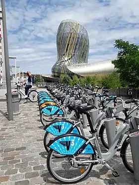 Photographie d'une station de VCub. Les vélos bleus et rouges sont alignés devant l'imposante cité du vin.