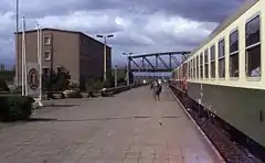 Photographie d'un train arrêté le long d'une plateforme ferroviaire à la fin de laquelle se trouve un pont en arc métallique. Un bâtiment en béton et un panneau portant les insignes de la RDA sont visibles à côté de la plateforme. Plusieurs personnes marchent sur la plateforme et les portes du train sont ouvertes.