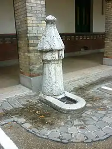 Fontaine à pompe : une borne en forme de pompe surmontant un puits déverse l'eau par un conduit dans une auge monolithique.