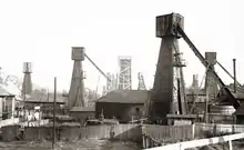 Photographie monochrome. Plusieurs puits de pétrole, en partie en bois ; à leur pied, des palissades en bois et quelques bâtiments.