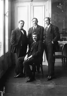 Photographie noir et blanc d'un homme assis entouré de trois hommes debout.