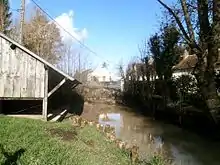 Photographie en couleurs d'un lavoir en bois établi sur une petite rivière ; un pont à l'arrière-plan.