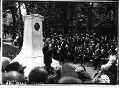Un homme fait un discours dans un parc public devant une foule assise.