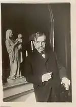 Paul Bourget, moustache, costume sombre, est adossé à une cheminée sur laquelle est exposée la statue d'une vierge à l'enfant. La main gauche du romancier est appuyée sur le dossier d'une chaise. Sa main droite semble tenir une cigarette.