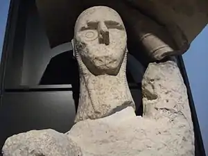Une statue partiellement reconstituée en pierre crème, accrochée à un portoir noir, le bouclier au-dessus de la tête.