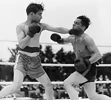 Photographie en noir et blanc de deux hommes équipés de gants et de shorts, se livrant un combat sur un ring de boxe.