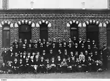 Photographie noir et blanc d'étudiants en quatre rangs avec leurs professeurs, devant un bâtiment en brique.