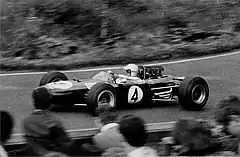 Photographie sépia d'une Brabham BT11, vue de profil, en piste, devant les têtes de plusieurs spectateurs, de dos.