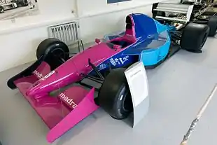 Photo d'une monoplace de Formule 1 de 1992 au nez rose, à l'arrière bleu et à l'aileron arrière blanc, dans un musée