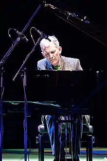 un homme assis au piano, en costume gris, cheveux gris