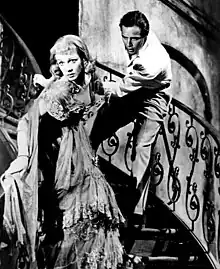 Photo en noir et blanc d'une femme vêtue d'une robe à froufrous, dans les escaliers, tentant d'échapper à un homme qui la retient par le bras.