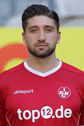 Photographie portrait d’un homme, portant une barbe de quelques jours, un maillot de football rouge sur les épaules