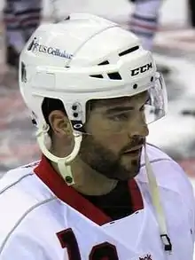 Photographie couleur d’un homme, barbu, de 3 quarts profil, portant un casque de hockey sur glace.