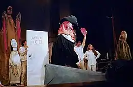 Des comédiens sur scène au milieu des marionnettes ; l'un d'eux tient une porte en carton portant l'inscription « LA PORTE DU CIEL » et un autre un cœur marqué GO TO HELL.