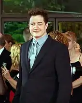 La photographie montre Brendan Fraser en plan américain. L'acteur a la peau claire, les yeux bleu-gris, les cheveux noirs. Il porte un costume.